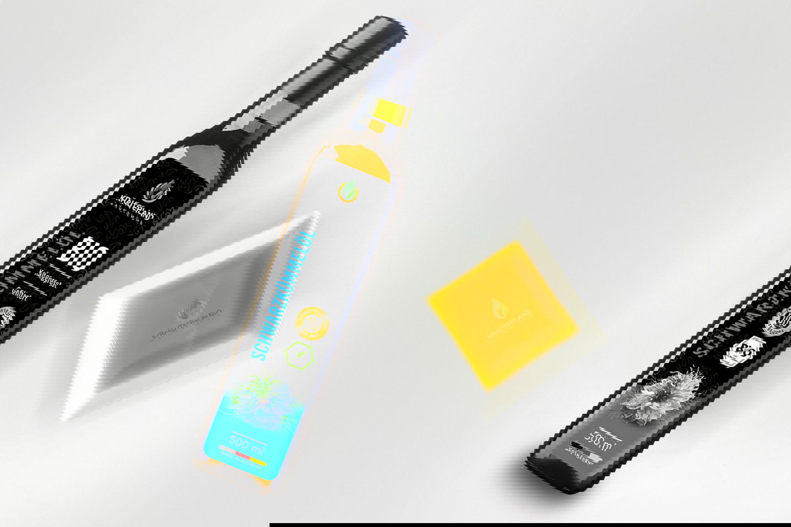 Kräuterland Bio Schwarzkümmelöl 500 ml kaltgepresst und gefiltert in Glasflasche neben Schälchen mit dem gleichen goldgelben Öl zur Veranschaulichung der Farbe