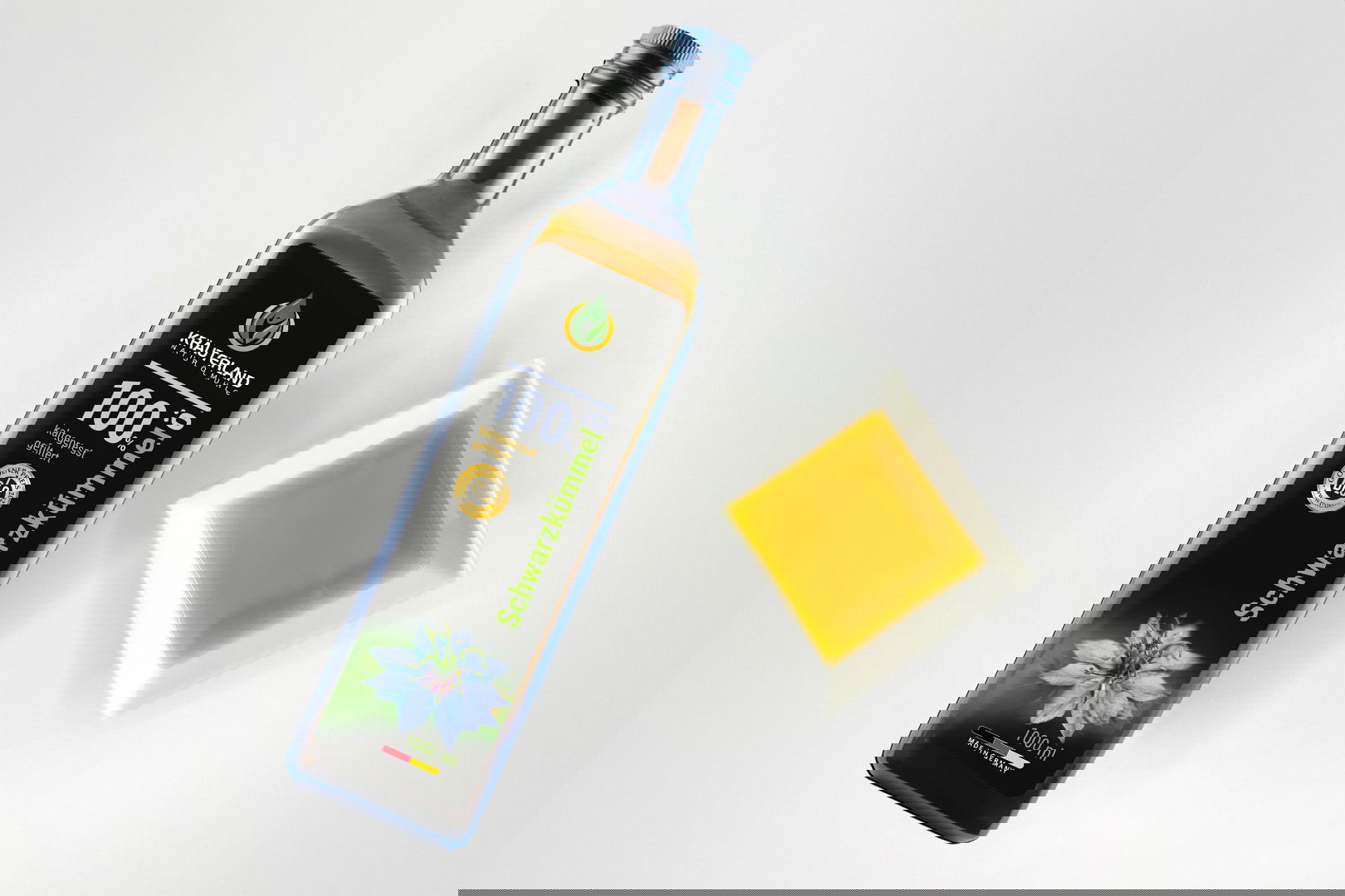 Kräuterland Schwarzkümmelöl 1000 ml kaltgepresst und gefiltert in Glasflasche neben Schälchen mit dem gleichen goldgelben Öl zur Veranschaulichung der Farbe