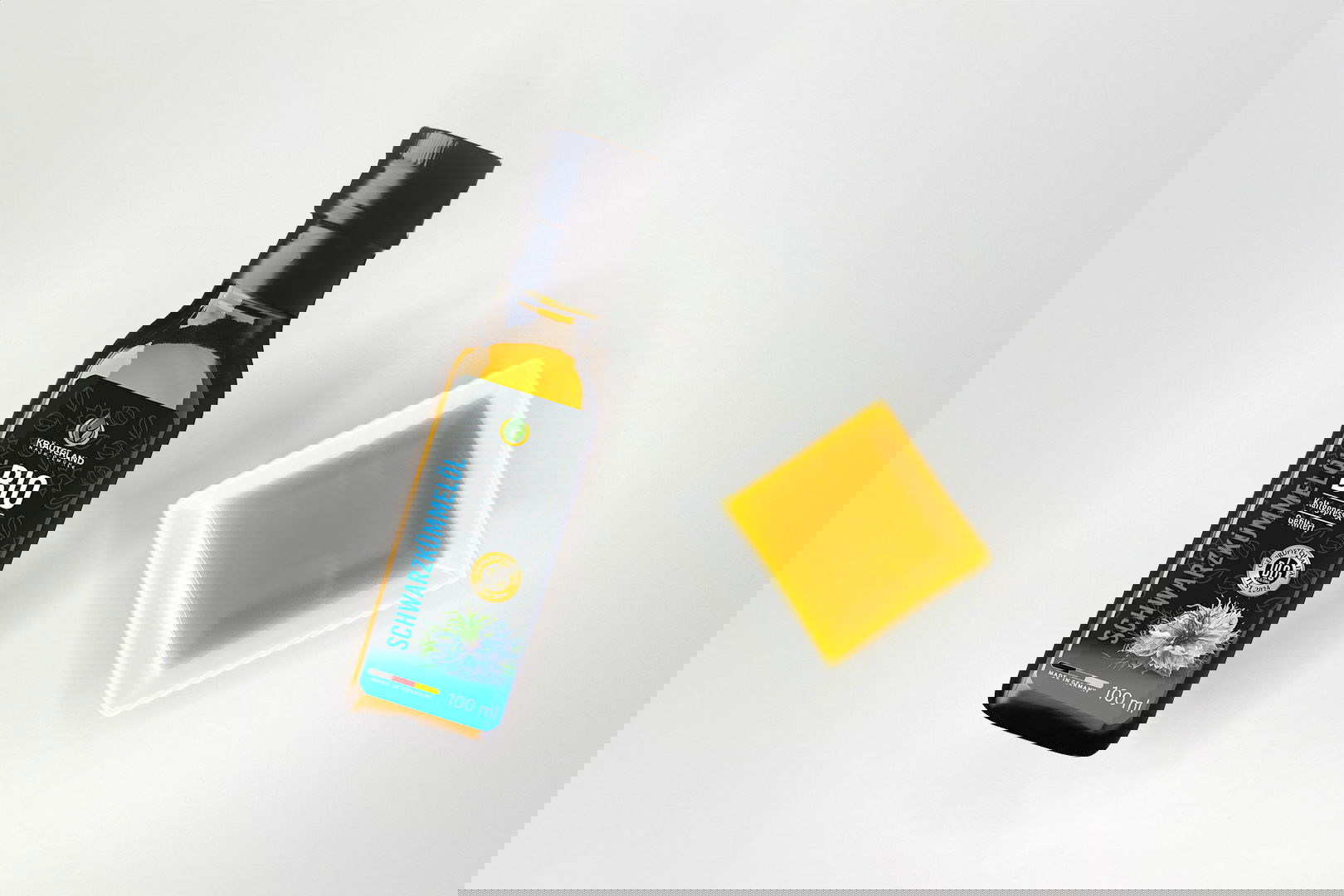 Kräuterland Bio Schwarzkümmelöl 100 ml kaltgepresst und gefiltert in Glasflasche neben Schälchen mit dem gleichen goldgelben Öl zur Veranschaulichung der Farbe